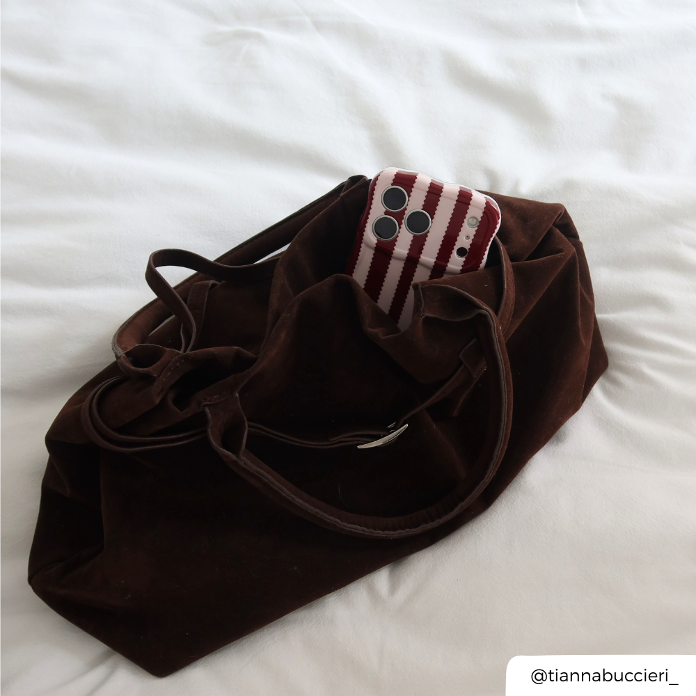 A dark brown velvet tote bag with drawstring straps, shown on white bedding with a phone in a burgundy and light pink striped wavy phone case peaking out the top of the bag.