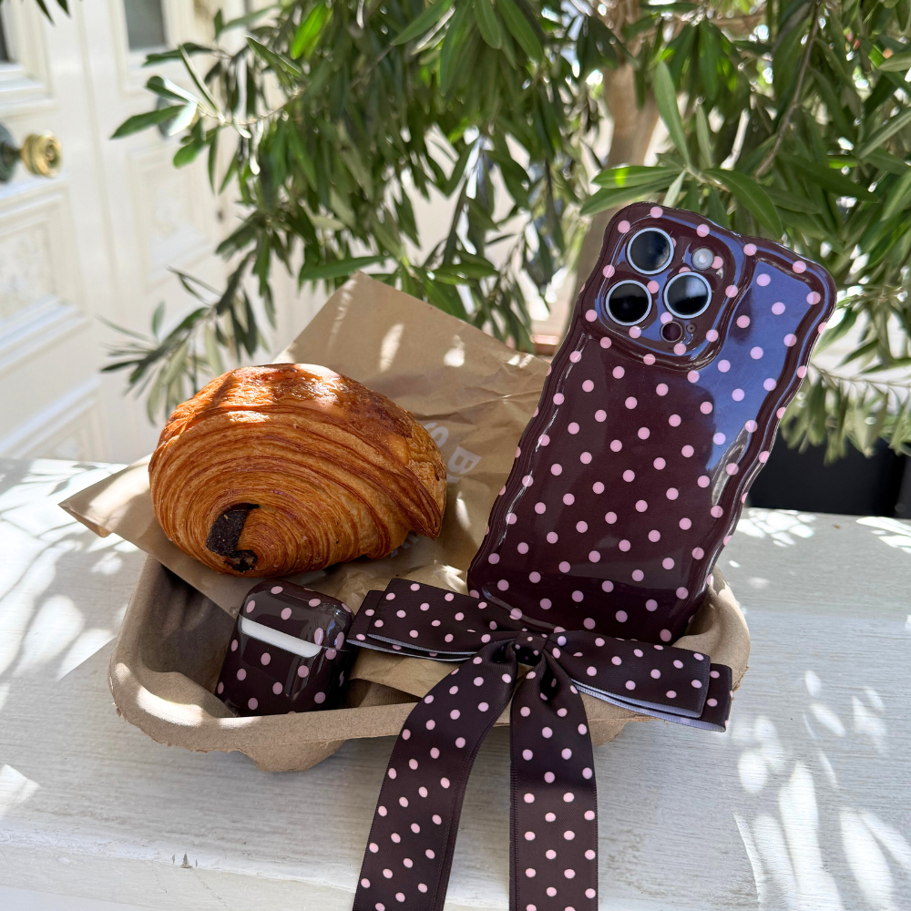 Chocolate croissant next to a brown polka dot Coconut Lane wavy phone case with ribbon, in sunlight by green leaves.