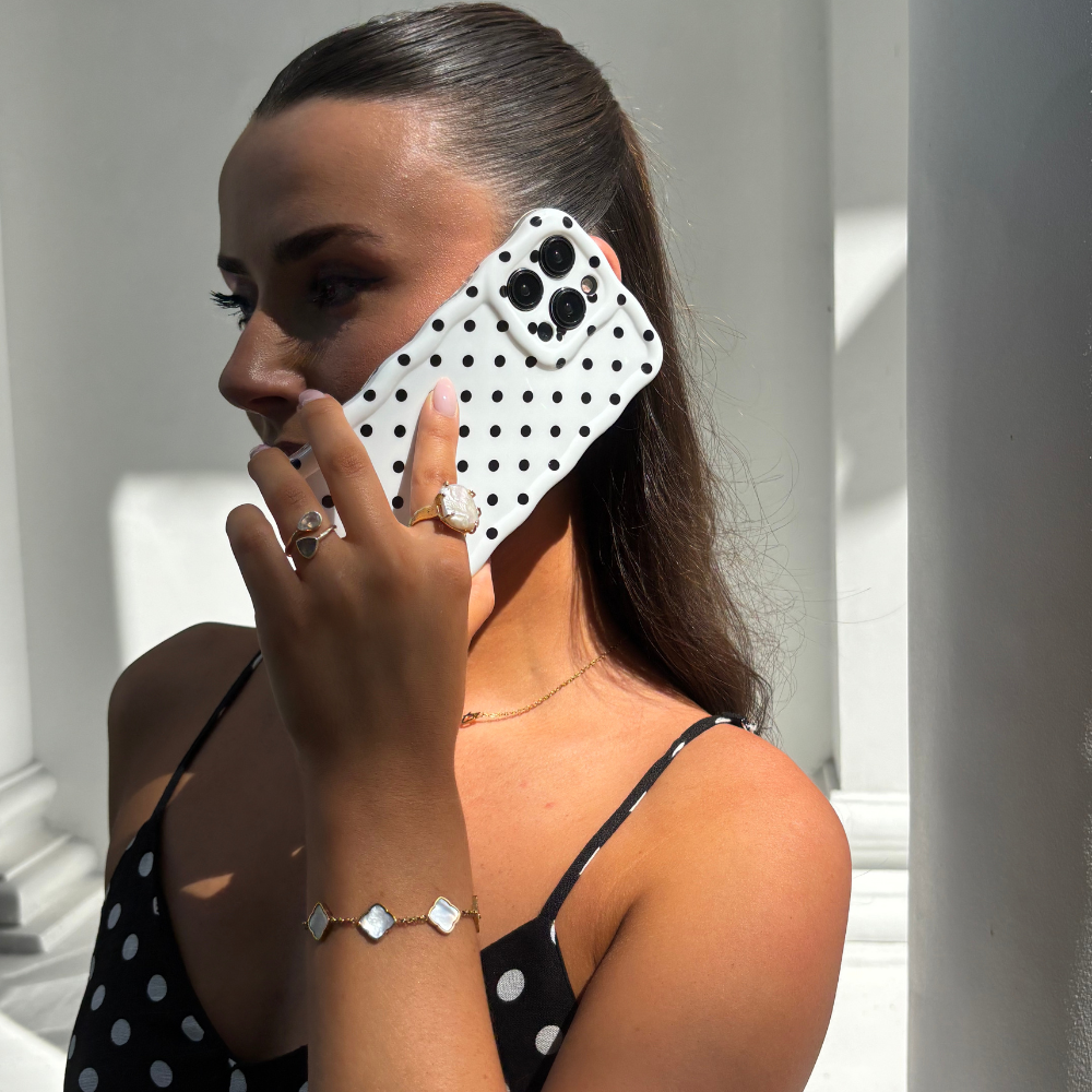 Woman in a black polka dot dress holds a white phone case with wavy edges and black polka dots to her ear.