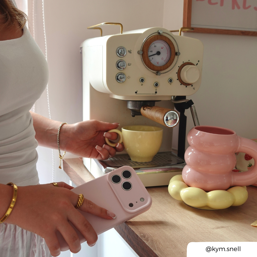a women making coffee in a soft, pastel kitchen setting, holding an iphone in a pastel pink phone case.