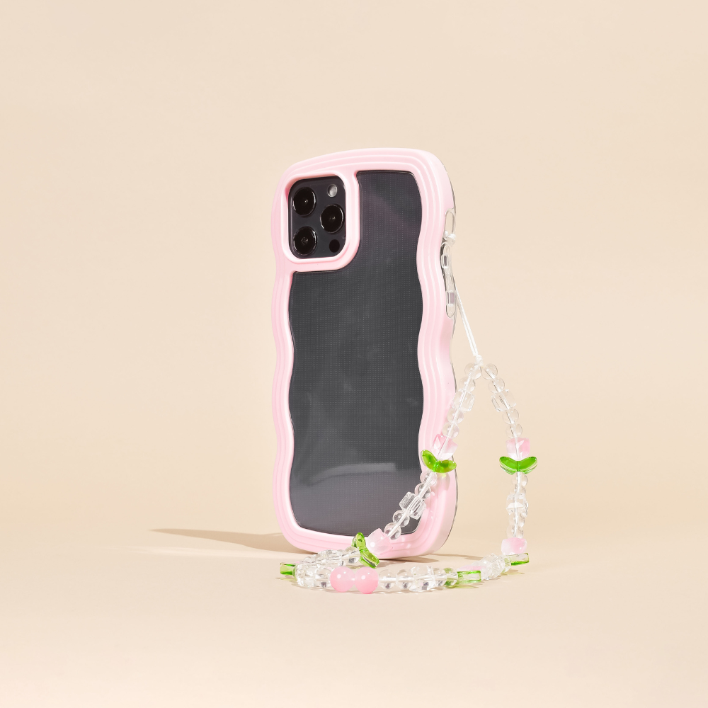 A smartphone case with a wavy pink edge, protecting a phone with triple rear cameras, is shown standing upright. Attached to the case is the Beaded Phone Strap - Cute Tulips by Coconut Lane, featuring small green leaves and pink beads that resemble cherries. This charming phone accessory stands against a plain beige background.