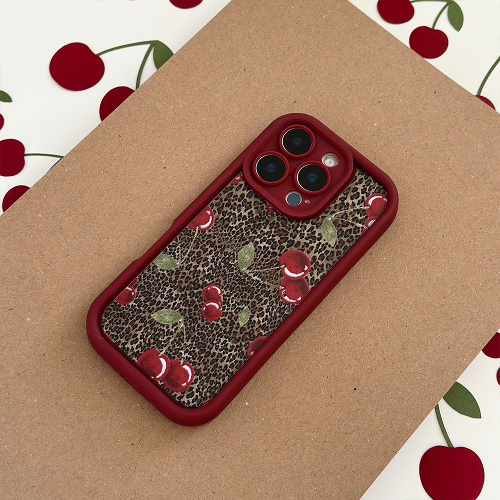A smartphone with a red case featuring a leopard print and cherry pattern rests on a brown surface, with cherry-themed paper in the background.