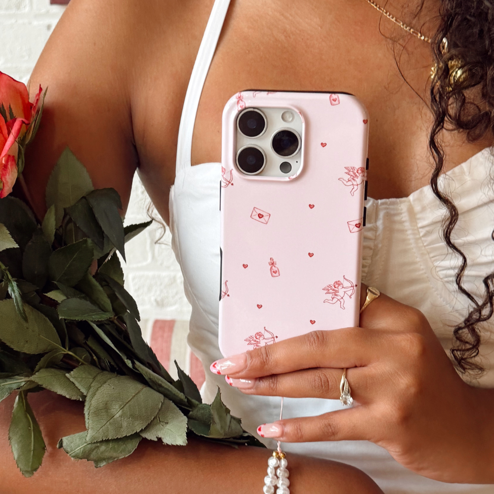 A hand with manicured nails holds an iPhone with a white case decorated with pink hearts and illustrations of pink cars carrying flowers.