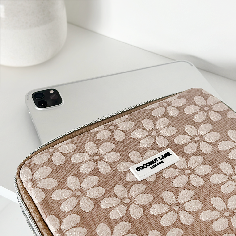 A beige floral-patterned laptop sleeve on a white surface with a tablet slightly protruding. A white ceramic vase is in the background.