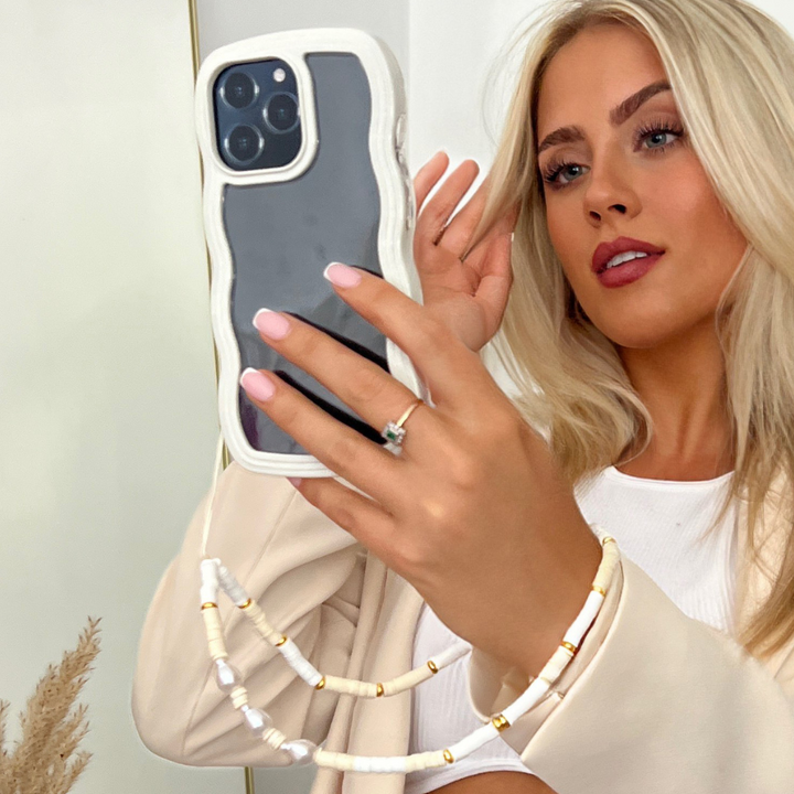 A blonde woman with long hair takes a mirror selfie using an iPhone 15 in a Curvy Phone Case - Cream by Coconut Lane, holding the phone with her left hand. She wears a light-colored jacket over a white top, showcasing a prominent ring on her ring finger and a decorative beaded phone strap.