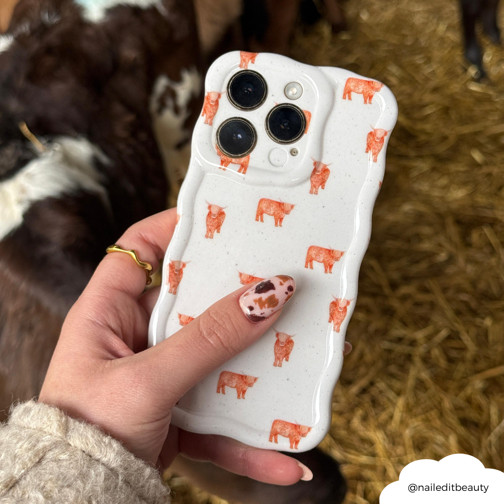 Hand with cow-print nails holding Coconut Lane wavy phone case featuring a highland cow pattern.