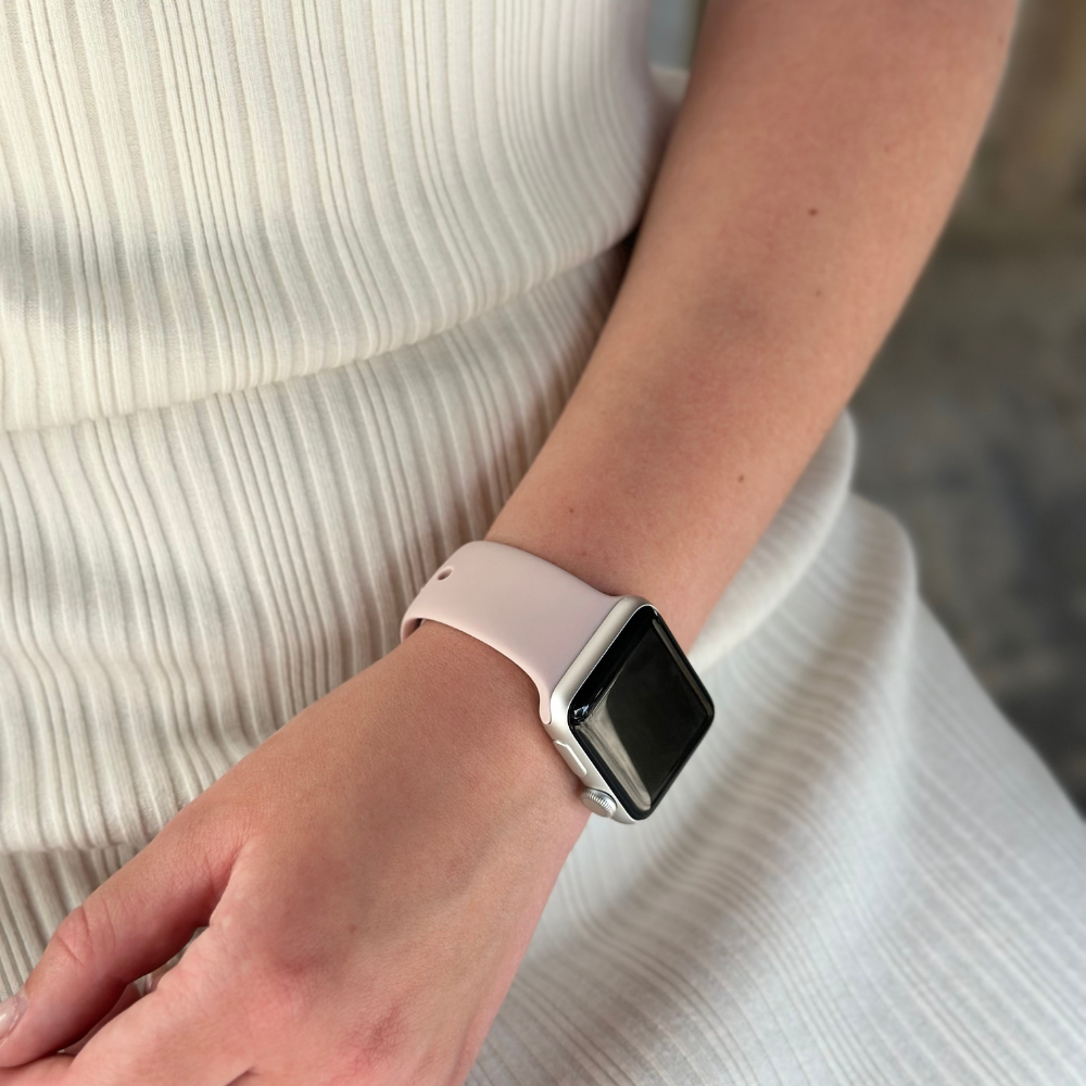 A person wearing a white textured dress is displaying their left wrist adorned with a sleek, modern smartwatch featuring a durable and chic Coconut Lane NAKD Apple Watch Strap in Dusty Pink. The background is blurred, drawing attention to the watch and the texture of the dress.