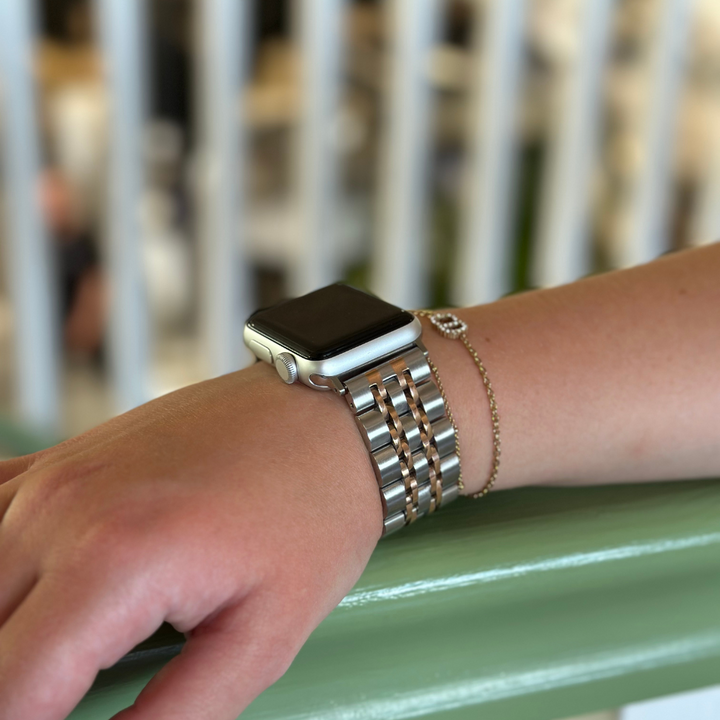 A close-up image features a person's wrist adorned with a Coconut Lane Stainless Steel Apple Watch Strap in Silver & Gold, paired with a delicate, thin gold-chain bracelet. The wrist rests on a green surface against a blurred white and beige background.