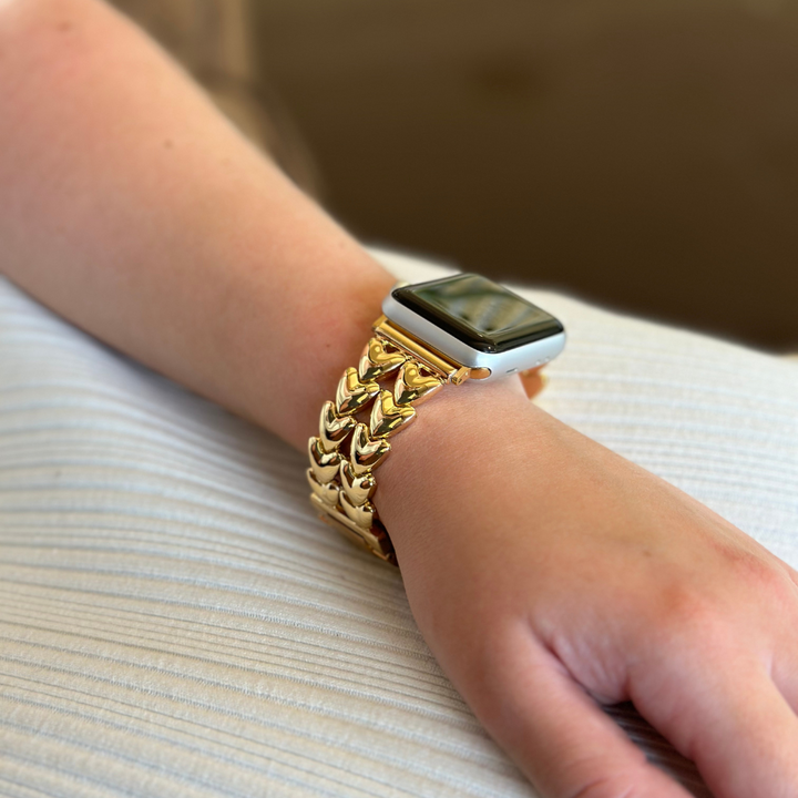 A close-up of a person's wrist shows them wearing an Apple Watch with the Hearts Chain Apple Watch Strap - Gold by Coconut Lane. The hand rests on a white, textured surface, creating a striking contrast with the gleaming, gold band and the sleek design of the smartwatch.