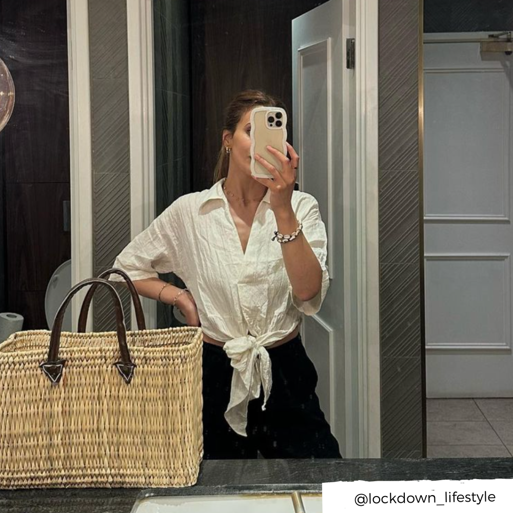 A person taking a mirror selfie in a bathroom, proudly showcasing their Curvy Phone Case - Cream from Coconut Lane on their iPhone 15. They are dressed in a white shirt tied at the waist and black pants, with a large woven straw bag resting on the counter. The Instagram handle @lockdown_lifestyle is visible in the bottom right corner.