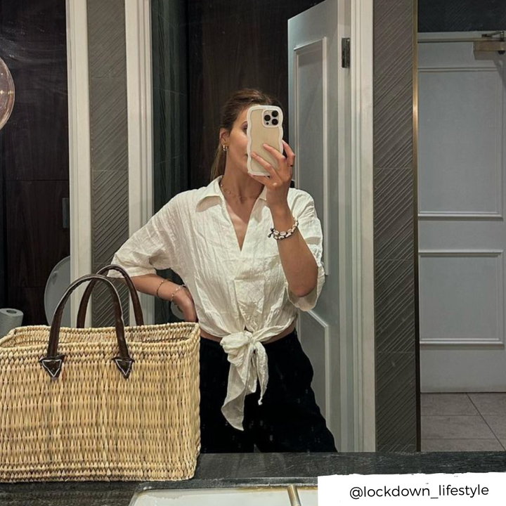 A person taking a mirror selfie in a bathroom, proudly showcasing their Curvy Phone Case - Cream from Coconut Lane on their iPhone 15. They are dressed in a white shirt tied at the waist and black pants, with a large woven straw bag resting on the counter. The Instagram handle @lockdown_lifestyle is visible in the bottom right corner.