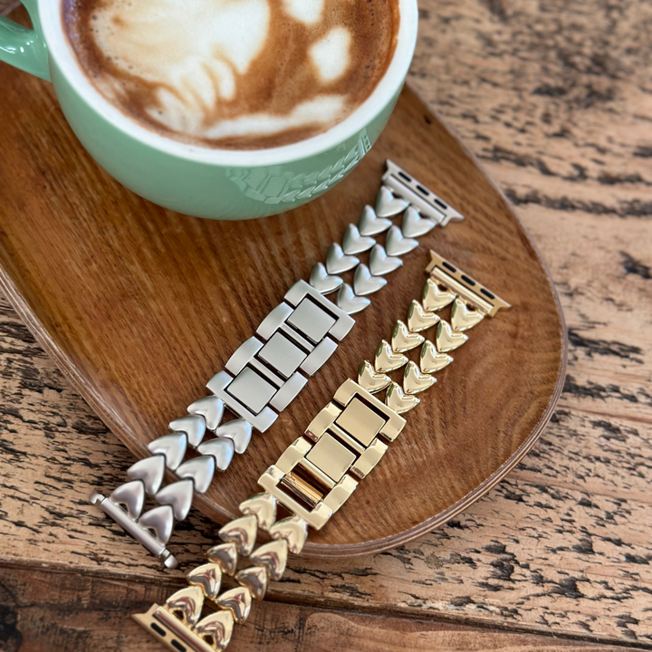 A wooden tray holding a green cup of cappuccino with latte art sits on a rustic wooden table. Next to the cup are two metallic watch bands, one silver and the Hearts Chain Apple Watch Strap - Gold from Coconut Lane, both featuring an adjustable stainless steel design perfect for various Apple Watch models.