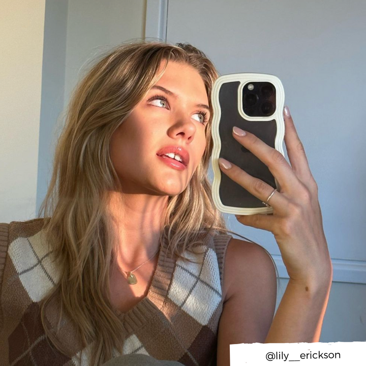 A person with long blonde hair takes a selfie in a well-lit room using a Curvy Phone Case - Cream from Coconut Lane. They are wearing a brown and white argyle sweater vest, gazing slightly upward. The Instagram handle "@lily__erickson" is visible at the bottom right.