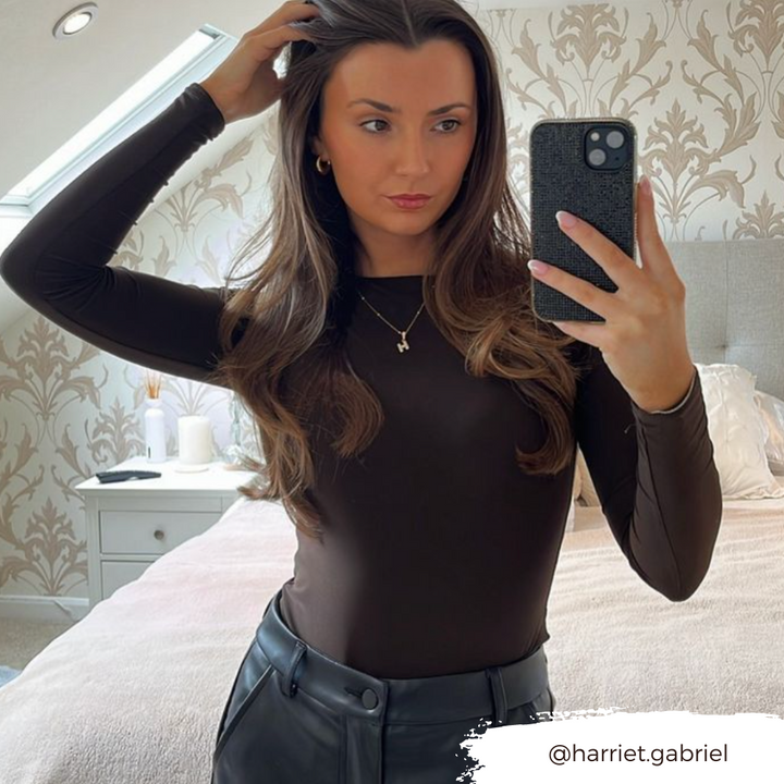 A person with long, wavy hair stands in a bedroom, wearing a dark brown top and black pants, taking a mirror selfie with their phone encased in Coconut Lane's extra protective Disco Sparkle Phone Case - Black. The room features wallpaper with a floral pattern, light-colored bedding on the bed, and a white nightstand. Instagram handle @harriet.gabriel.