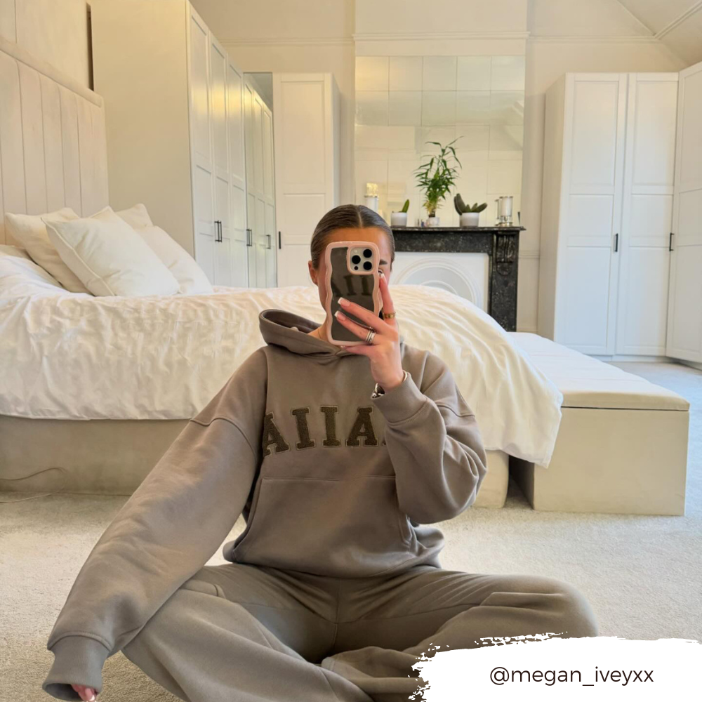 A person wearing a gray hooded sweatshirt and matching sweatpants is sitting cross-legged on a carpeted floor in front of a bed. They are taking a mirror selfie with their phone, which features the Coconut Lane Curvy Mirror Phone Case - Pink. An Instagram handle, @megan_iveyxx, is displayed in the bottom right corner.