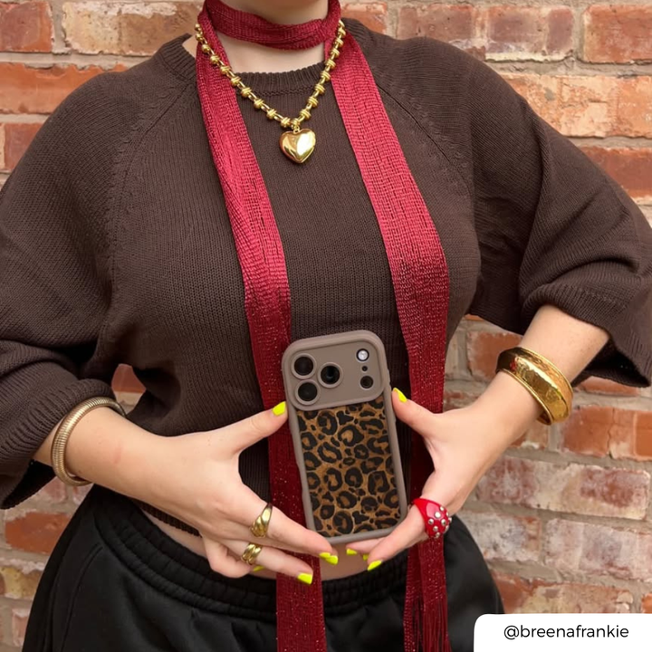 A woman wearing a chocolate brown jumper, accessorised with gold jewellery, a red mesh scarf. She is holding an iphone in a mocha leopard print phone case Her nails are painted yellow. 
