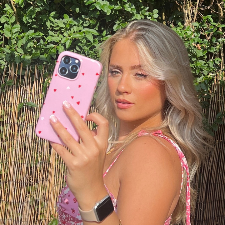 A woman with long blonde hair takes a selfie with a pink phone adorned with the "Cute Hearts Phone Case" by Coconut Lane, likely for an iPhone15. She is wearing a pink floral top and a smartwatch, standing outdoors among leafy green plants and beside a bamboo fence. The sunlight highlights her face beautifully, making it perfect for Valentine's Day.