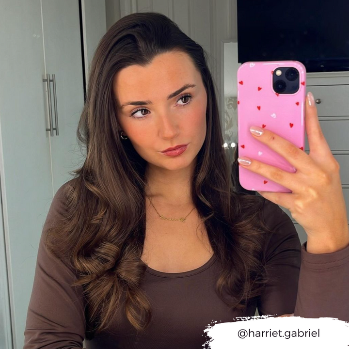 A woman with long, wavy brown hair is taking a mirror selfie with her iPhone, which is protected by the Cute Hearts Phone Case from Coconut Lane. She is wearing a brown top and a delicate necklace. The Instagram handle "@harriet.gabriel" is written in the bottom right corner.