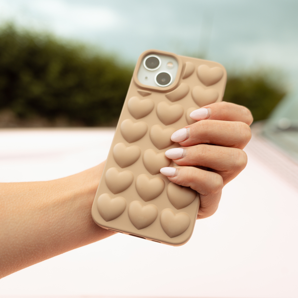 A hand with manicured nails holds an iPhone 15 adorned in a Coconut Lane's "3D Hearts Phone Case - Matte Nude," featuring a beige heart pattern. The matte finish adds elegance, while the background is slightly blurred, showcasing greenery and a light-colored surface.