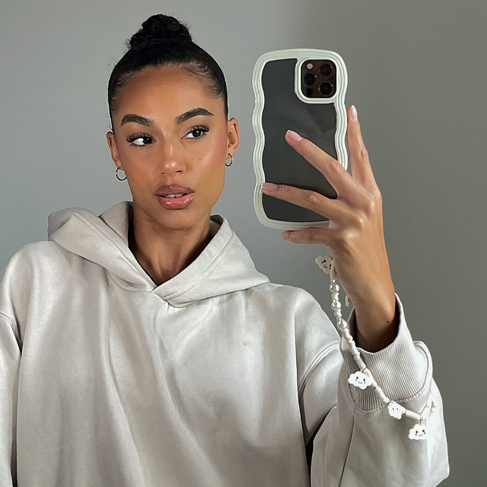 A person with their hair in a bun takes a mirror selfie. They are wearing a light-colored hooded sweatshirt and holding an iPhone 15 with Coconut Lane's Curvy Phone Case in Cream, which is highly protective and has a decorative charm. The background is plain and neutral.