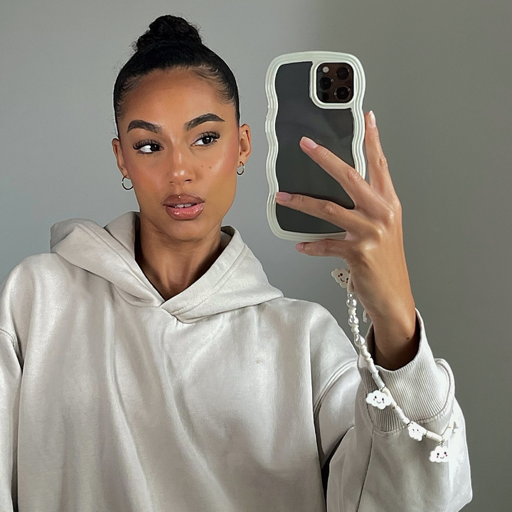 A person with their hair in a bun takes a mirror selfie. They are wearing a light-colored hooded sweatshirt and holding an iPhone 15 with Coconut Lane's Curvy Phone Case in Cream, which is highly protective and has a decorative charm. The background is plain and neutral.