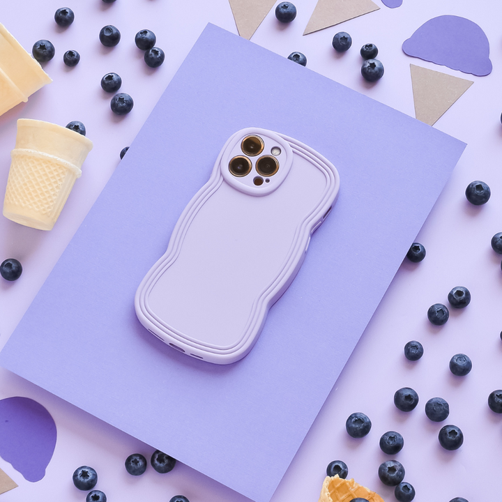 A smartphone in a lightweight, blue NAKD Curvy Phone Case by Coconut Lane rests atop a light purple sheet on a flat surface. Scattered around are blueberries, pieces of waffle cones, and cut-out shapes of ice cream cones and scoops in purple, brown, and white. The scene has a playful and colorful aesthetic.