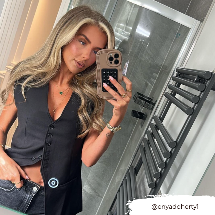 A woman with long blonde hair stands in a modern bathroom taking a mirror selfie with her phone encased in the NAKD Curvy Phone Case - Coffee by Coconut Lane. She is wearing a black sleeveless top and dark jeans. Behind her is a gray tiled wall and a black towel radiator. The Instagram handle @enyadoherty1 is in the bottom right corner.