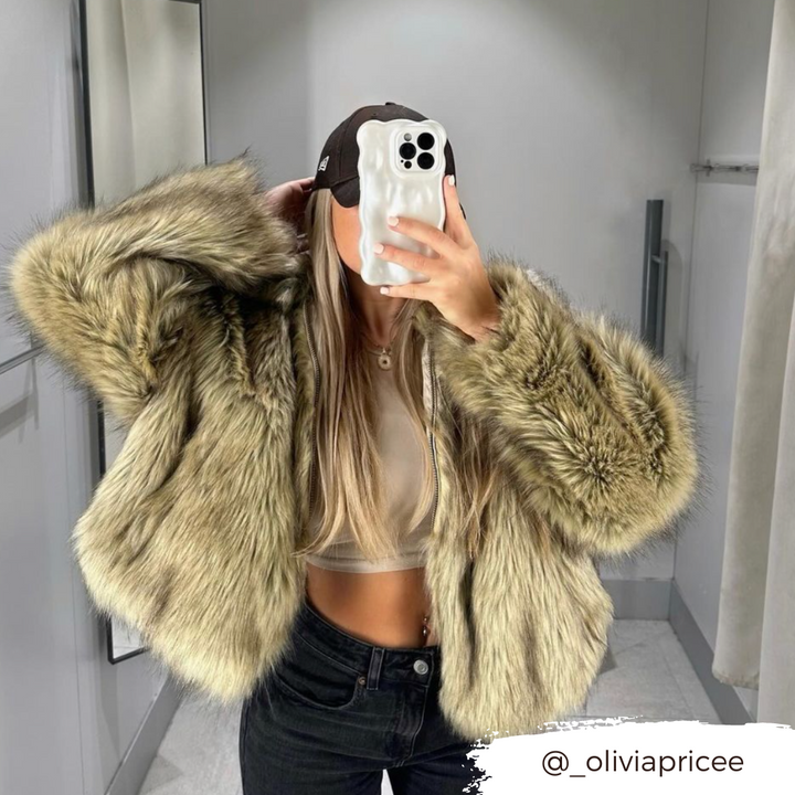 A person takes a mirror selfie in a well-lit room. They are wearing a tan crop top and black pants, topped with a large, fluffy, tan fur coat. They also have long, blonde hair and a black baseball cap. The Instagram handle @_oliviapricee is visible in the corner along with their 3D Pearl Phone Case - White from Coconut Lane.