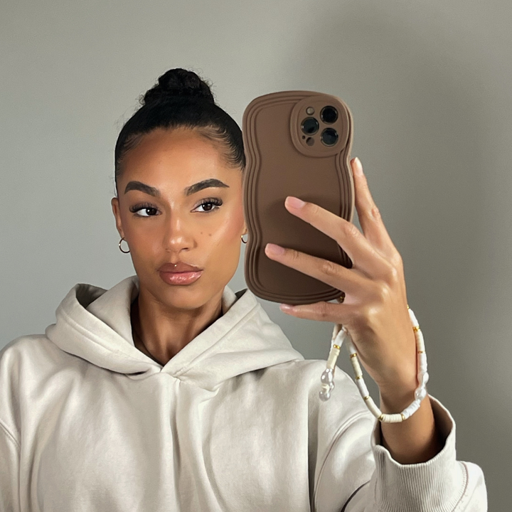 A woman with her hair in a high bun takes a selfie in front of a mirror. She is wearing a light beige hoodie and holding a brown phone with the lightweight, highly protective NAKD Curvy Phone Case in "Coffee" by Coconut Lane, featuring wavy edges. A beaded charm is attached to her phone case. The background is plain and gray.