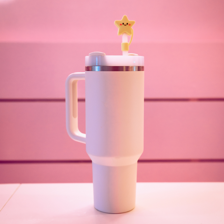 A tall, pure white insulated tumbler with a generous handle is set on a surface against a pale pink backdrop. The tumbler is topped with Coconut Lane's Star Straw Cover, which includes a yellow star-shaped straw topper that has a smiling face.