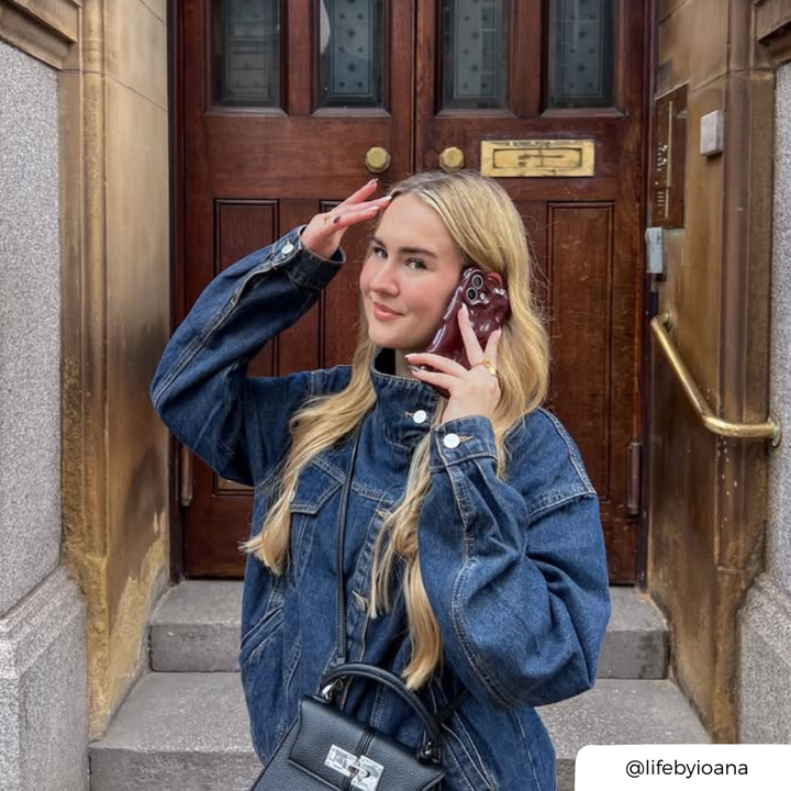 A woman with long blonde hair, wearing a dark blue denim jacket in front of a brown door holds an iphone up to her ear. The phone is in a 3d bubble burgundy phone case.