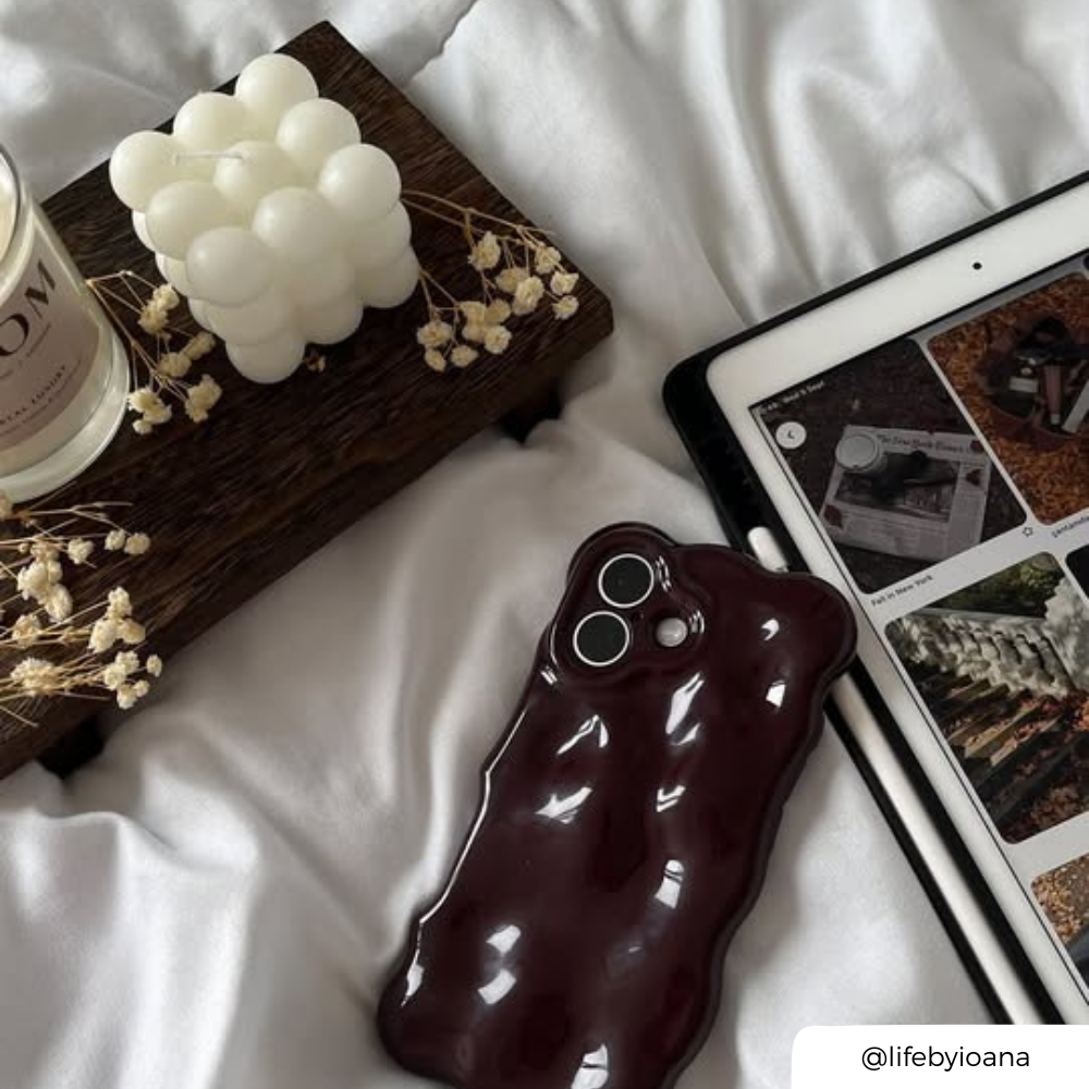 A glossy, wavy-edged phone case in a deep burgundy colour, and 3d bubble effect, shown on a white fabric surface next to a tablet, a candle, and dried flowers.