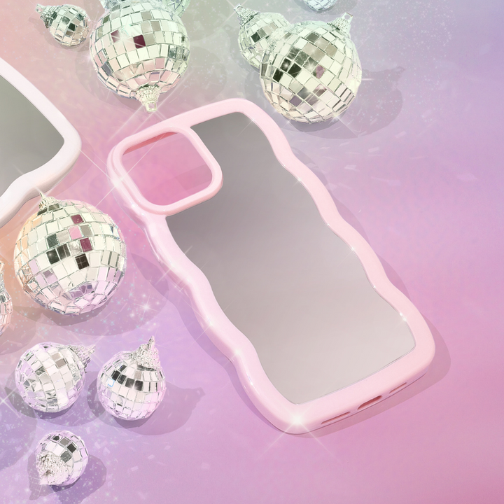 A pink, wavy-edge phone case lies on a pastel, shimmering background. Surrounding the Curvy Mirror Phone Case - Pink by Coconut Lane are small, reflective disco ball ornaments, adding a festive and sparkly touch to the scene. The background blends hues of purple and pink, accentuating the glittery ambiance.