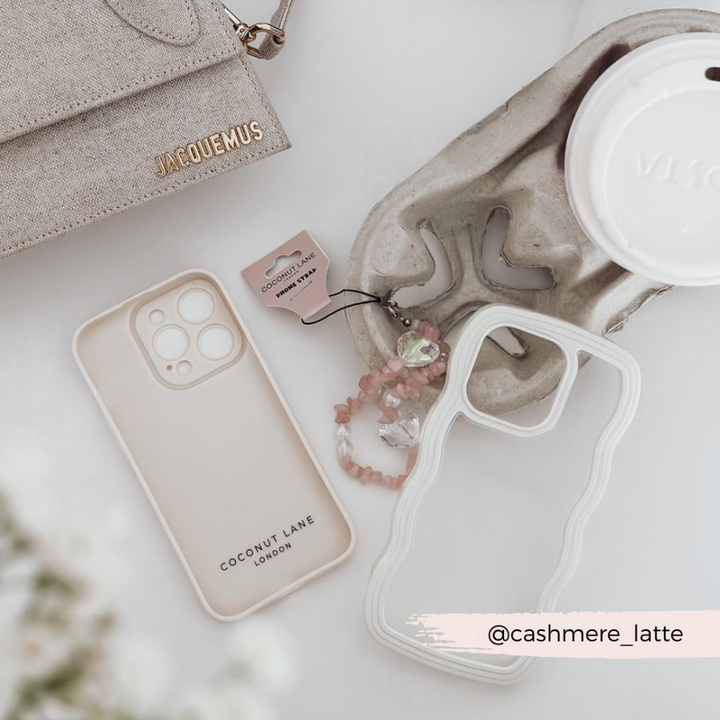 A stylish flat lay featuring a beige Jacquemus bag, a white coffee cup with a lid, two highly protective iPhone 15 cases (one beige and one Coconut Lane Curvy Phone Case in cream), pink beaded accessories, and a tag labeled "Coconut Lane London". The handle @cashmere_latte is displayed on the bottom right.
