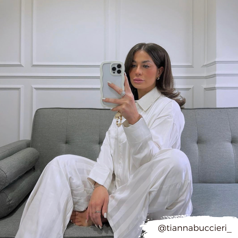 A woman with long dark hair is sitting on a gray couch taking a mirror selfie with her phone. She is wearing a white striped pajama set, and her Curvy Phone Case - Cream from Coconut Lane shines in the shot. The background features a white paneled wall. The Instagram handle "@tiannabuccieri_" is displayed in the corner.