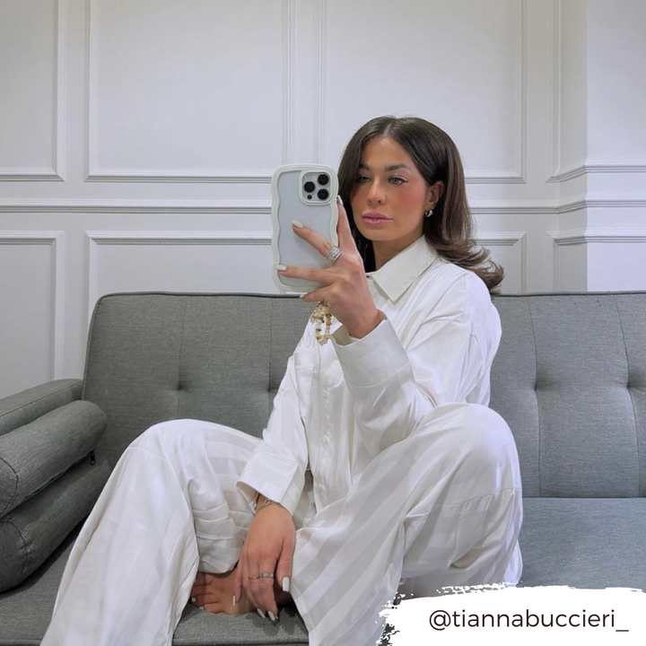 A woman with long dark hair is sitting on a gray couch taking a mirror selfie with her phone. She is wearing a white striped pajama set, and her Curvy Phone Case - Cream from Coconut Lane shines in the shot. The background features a white paneled wall. The Instagram handle "@tiannabuccieri_" is displayed in the corner.
