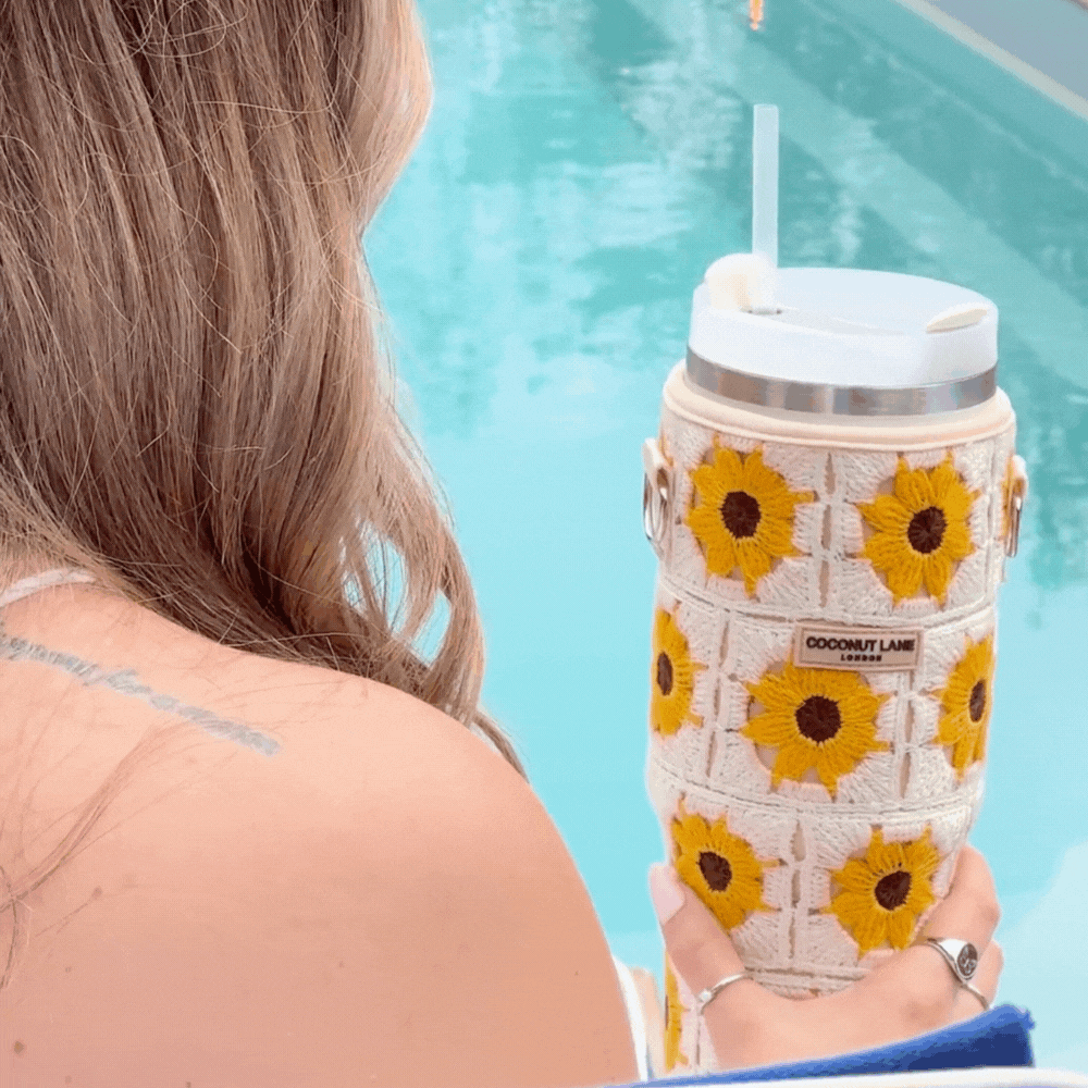 A person with long, wavy hair, seen from behind, holds a Coconut Lane Tumbler Carry Case - Sunflower Crochet. The person is by a pool, with one shoulder visible, wearing a sleeveless top. The water is bright blue and the overall scene has a relaxed, summery vibe.