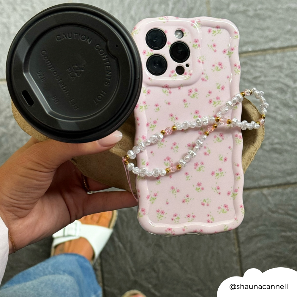 Hand holding Coconut Lane Wavy Phone Case in pink ditsy floral with beaded strap; white shoes and blue jeans in background.