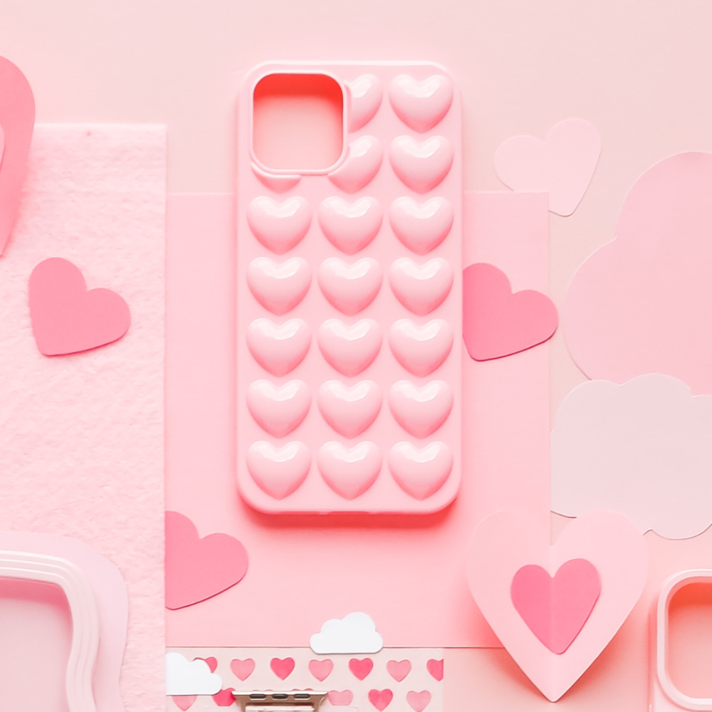 A **Coconut Lane 3D Hearts Phone Case - Candyfloss Pink**, featuring a textured pattern of small, raised hearts, is artfully displayed amidst an array of pink and paper hearts and cloud shapes on a flat, pink surface. The entire composition exudes a theme of love and romance.