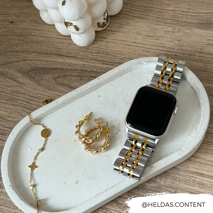 A Stainless Steel Apple Watch Strap - Silver & Gold by Coconut Lane with an adjustable strap rests on a decorative white tray, accompanied by gold earrings and a bracelet with a star charm. The tray sits on a wooden surface, featuring the Instagram handle @heldas.content in the bottom right corner.