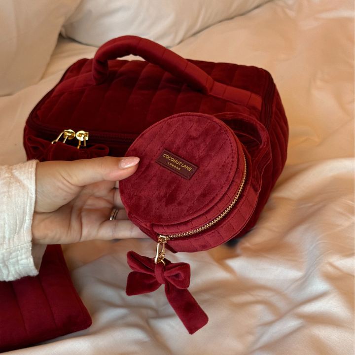 Burgundy velvet jewellery box with a quilted design, top handle, bow detail, and gold-tone hardware on a white bed