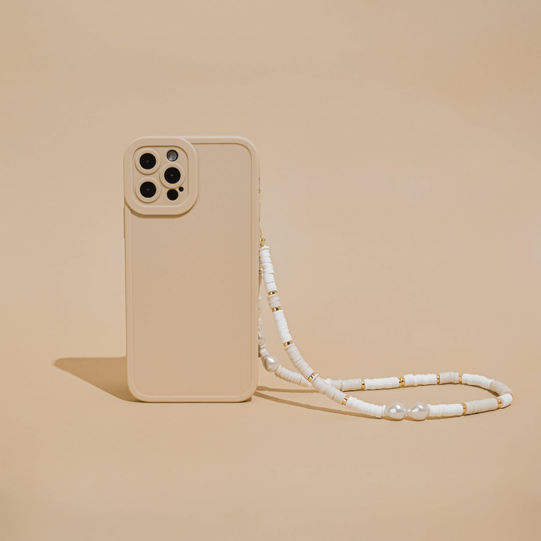 A Beaded Phone Strap in Nude & Pearl, created by Coconut Lane, adorns a beige phone case against a matching beige background. The case features a cutout for four camera lenses and is elegantly detailed with charming phone charms, complemented by white beads and small gold accents on the strap.