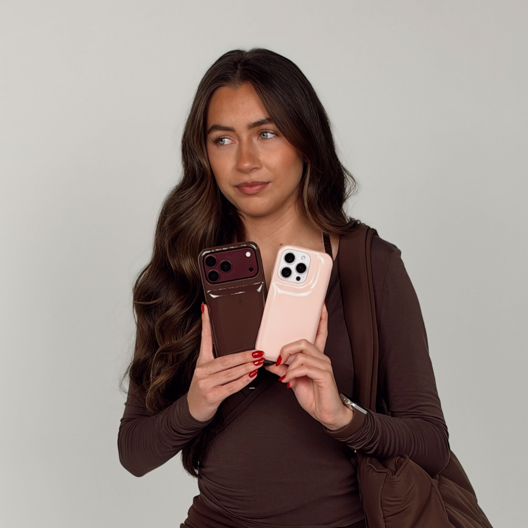 Woman wearing a brown pilates outfit holding two iphones. One is in a glossy chocolate brown Coconut Lane puffer phone case with embossed logo. The other iphone is in a pastel pink Coconut Lane puffer phone case.On her wrist is a Coconut Lane NAKD Chocolate brown apple watch strap. 