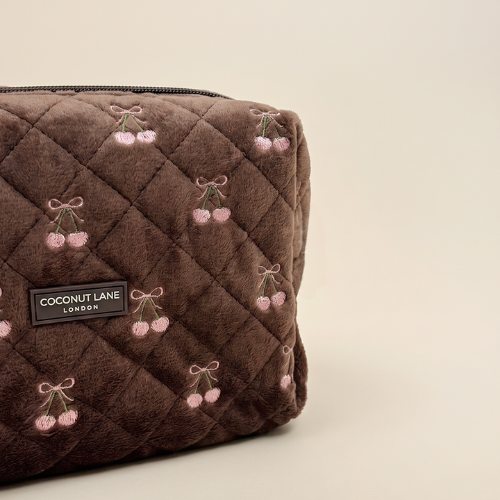 Close-up of a brown quilted velvet makeup bag with pink embroidered cherries and a “Coconut Lane London” label.