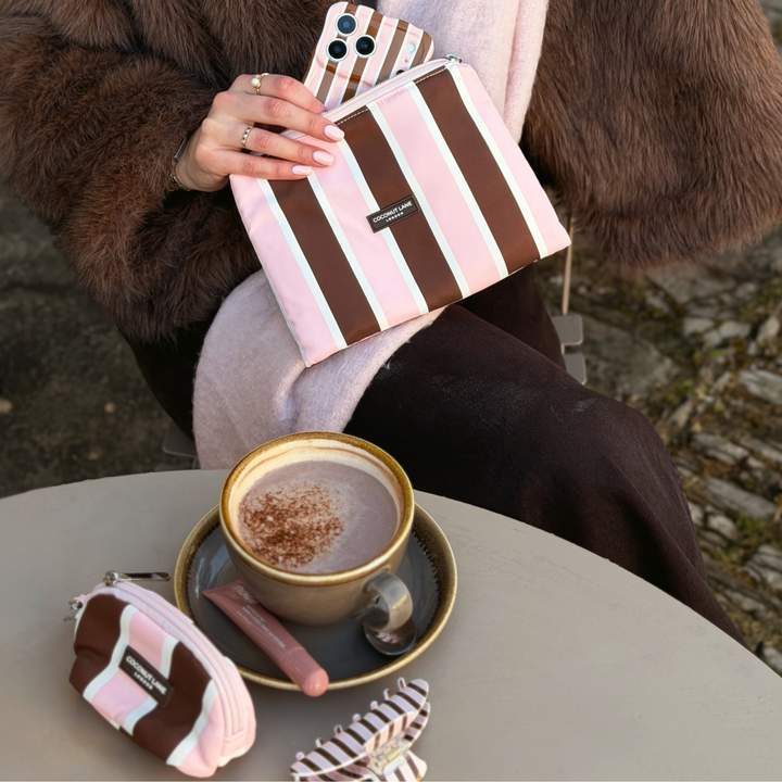Woman holding the Coconut Lane Cocoa & Pink Stripe Everything Pouch featuring chocolate brown, pastel pink and white vertical stripes. On her wrist is the matching satin scrunchie. She is wearing a baby pink scarf and a faux fur brown jacket. On the table is a coffee and a matching mini keyring and claw clip.