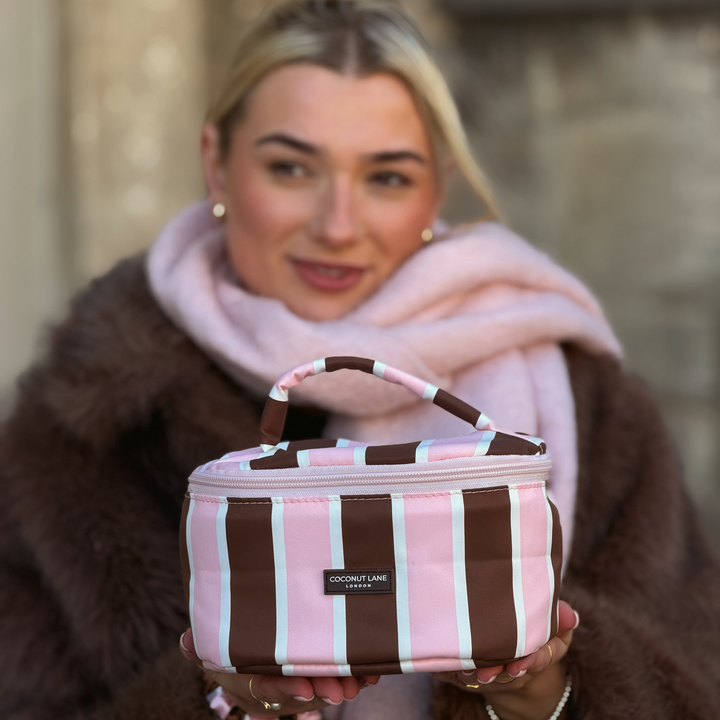 Woman with blonde hair, wearing a baby pink scarf and brown faux  fur jacket, holding a coconut Lane Cocoa & Pink Stripe round vanity makeup bag with chocolate brown and baby pink stripes.