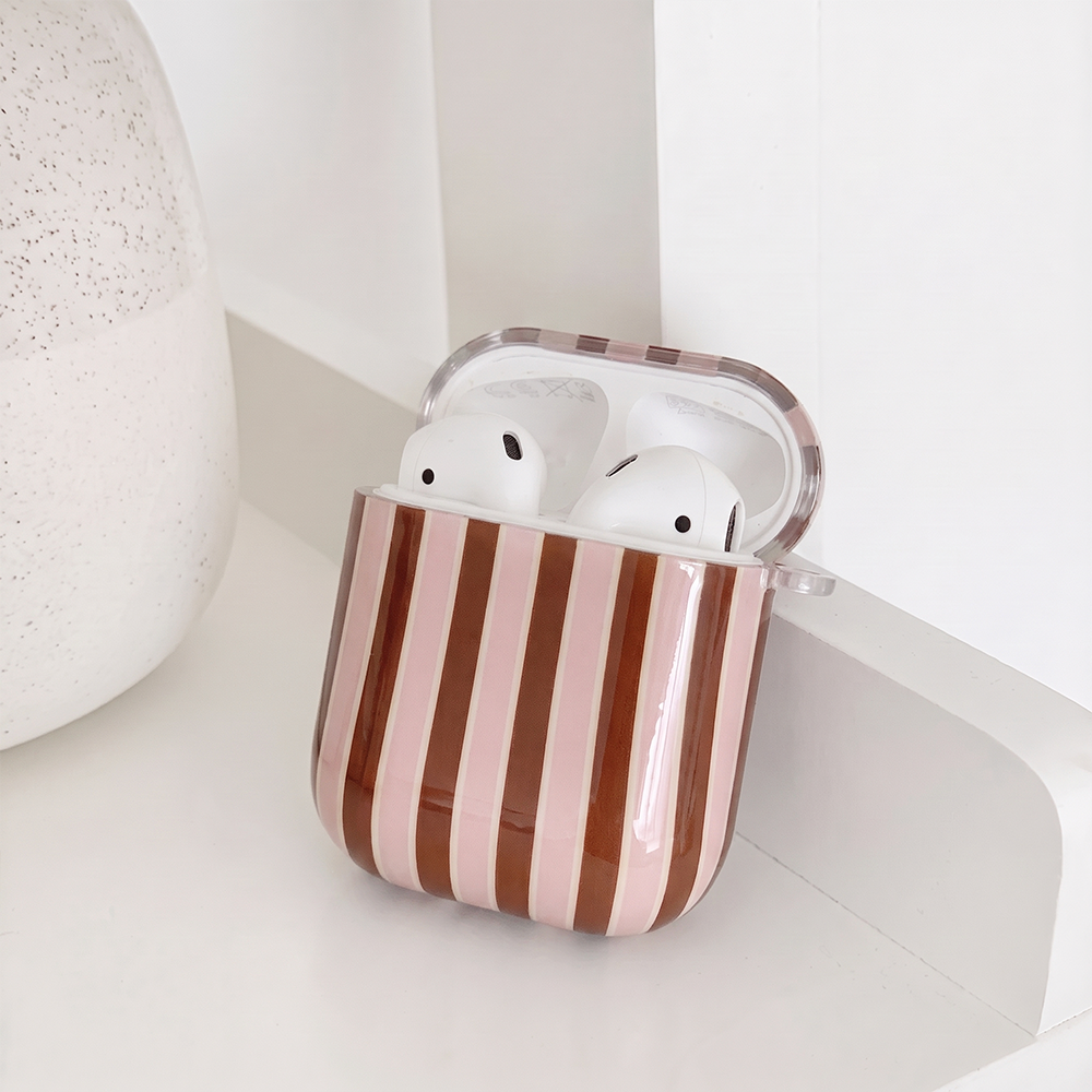 Showing the inside of a glossy AirPods Case cover with vertical stripes in pink and brown, separated by thin white lines, with a small clear clip loop on the side. It sits on a white shelf beside a white speckled vase.