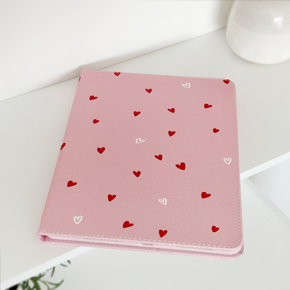 A Cute Hearts iPad Case by Coconut Lane, with a pink cover decorated with small red and white hearts, is placed on a white surface. The background features a white, blurred, round object that appears to be a ceramic pot, enhancing the minimalist aesthetic.