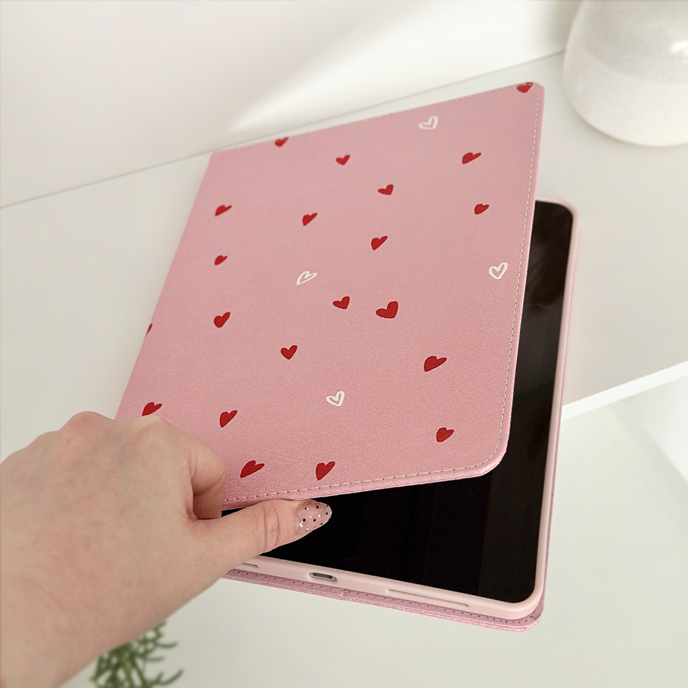 A hand is shown opening a Coconut Lane Cute Hearts iPad Case made from pink PU Vegan Leather and adorned with small red and white hearts. The device inside has a black screen, and the setting appears to be a light-colored, minimalist room with a small plant visible.