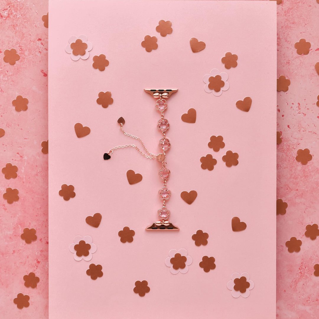 The Coconut Lane Rose Gold Flower Bracelet Apple Watch Strap is elegantly displayed flat against a pink background. Scattered around the strap are gold and white paper cutouts of hearts and flowers, crafting a romantic and sophisticated composition.